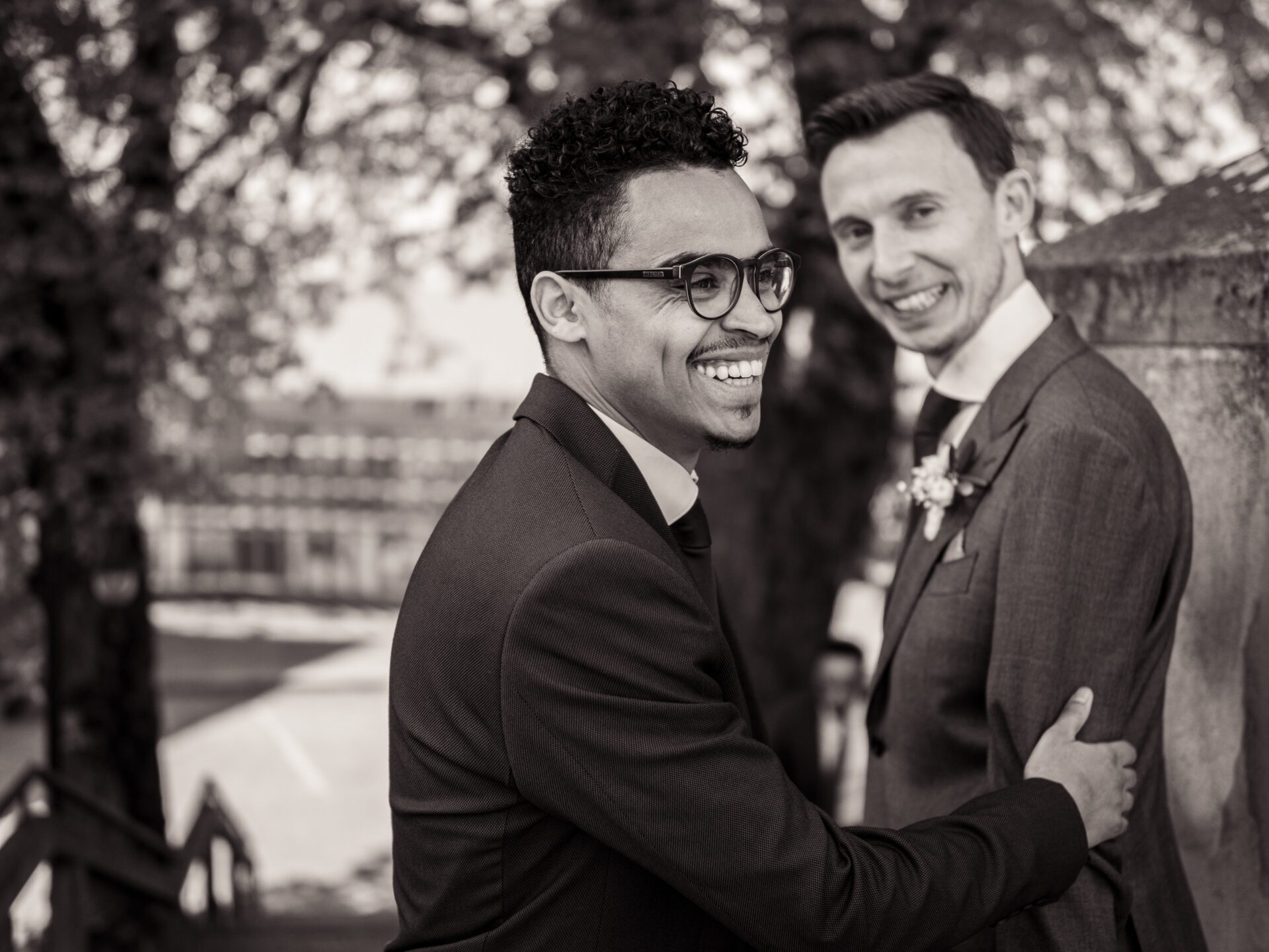 Zwei Männer in Anzügen lächeln sich im Freien freundlich an, der eine mit Brille und lockigem Haar stützt seine Hand auf den Arm des anderen. Im Hintergrund erscheinen Bäume und ein unscharfes Gebäude. Ein zeitloses schwarz-weiß Hochzeitsfoto von einem Hochzeitsfotograf in München. Hochzeitsfotos_Hochzeitsfotograf_München