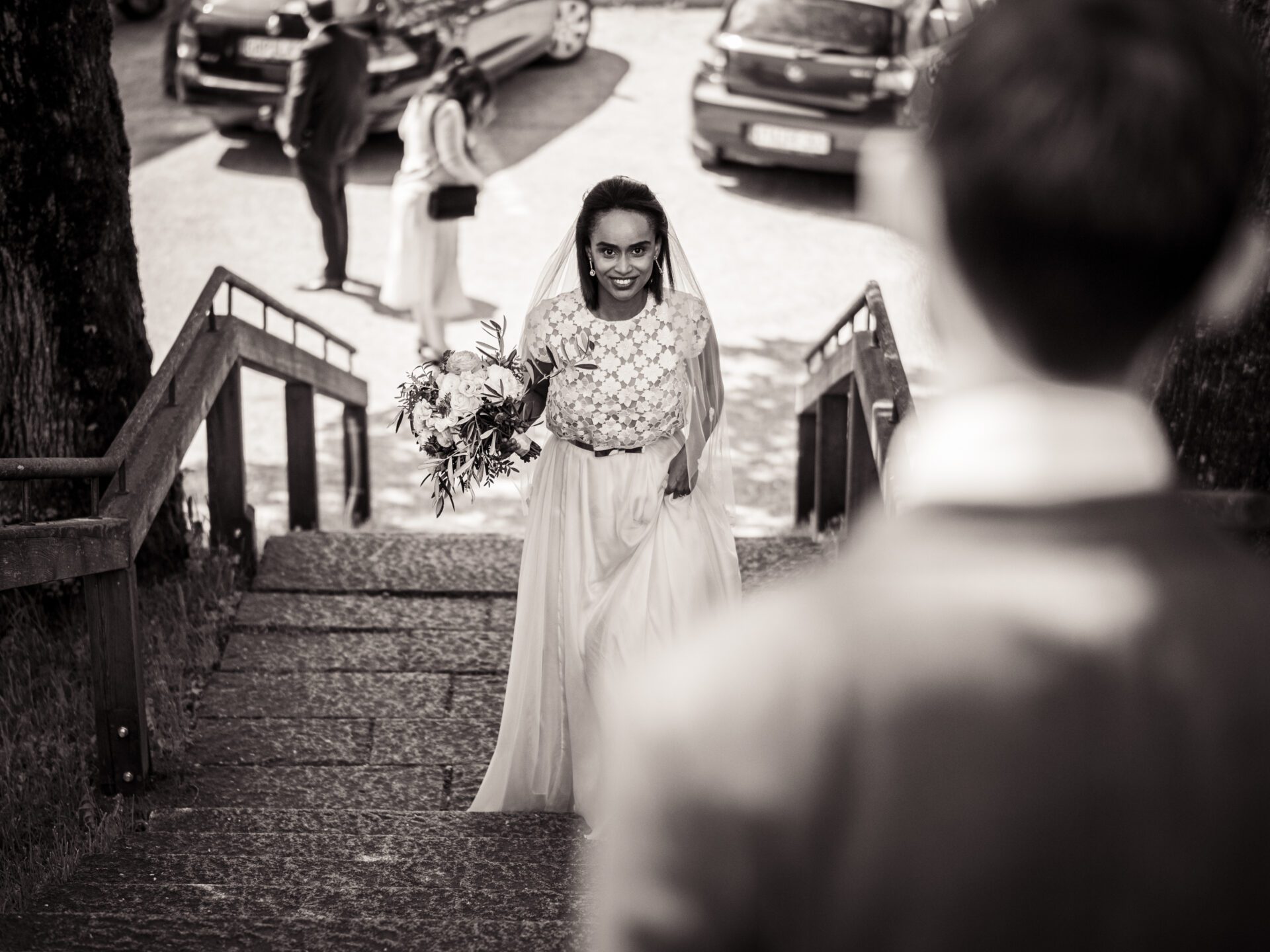 Eine Braut in Brautkleid und Schleier, die einen Blumenstrauß in der Hand hält, geht über eine Steintreppe auf eine Person im Anzug zu. Hinter ihnen sind geparkte Autos und Menschen zu sehen. Dieses schwarz-weiße Hochzeitsfoto fängt einen authentischen Moment in München ein. Hochzeitsfotos_Hochzeitsfotograf_München