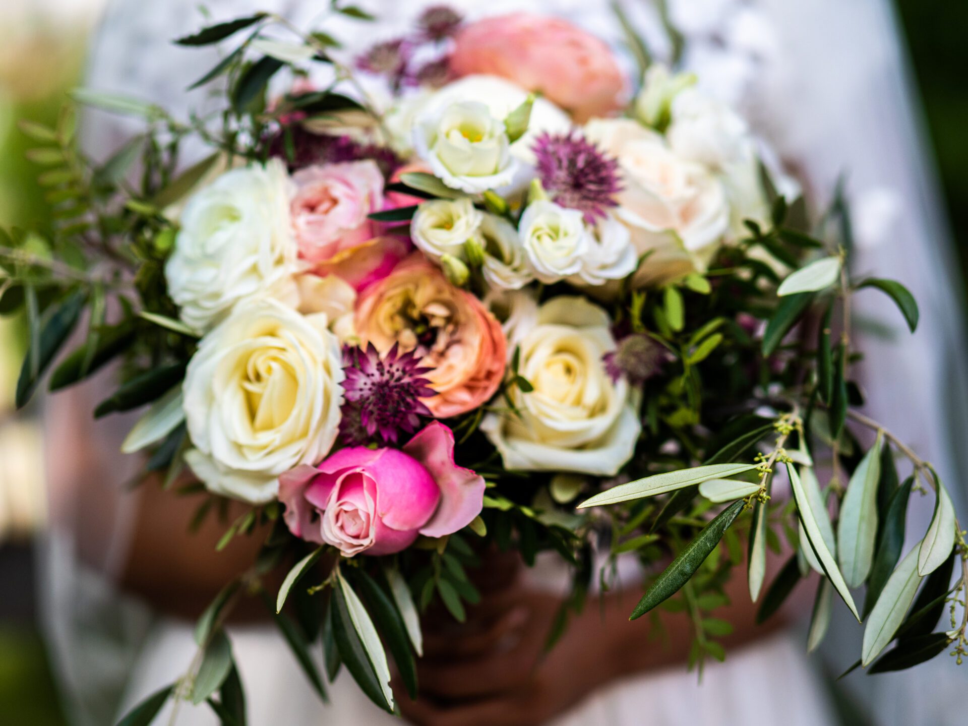 Eine Nahaufnahme eines farbenfrohen Brautstraußes mit weißen, rosafarbenen und pfirsichfarbenen Rosen, lila Distelblüten und grünem Laub, wunderschön eingefangen von einem Hochzeitsfotografen in München. Die perfekte Inspiration für Ihre Hochzeitsfotos. Hochzeitsfotos_Hochzeitsfotograf_München