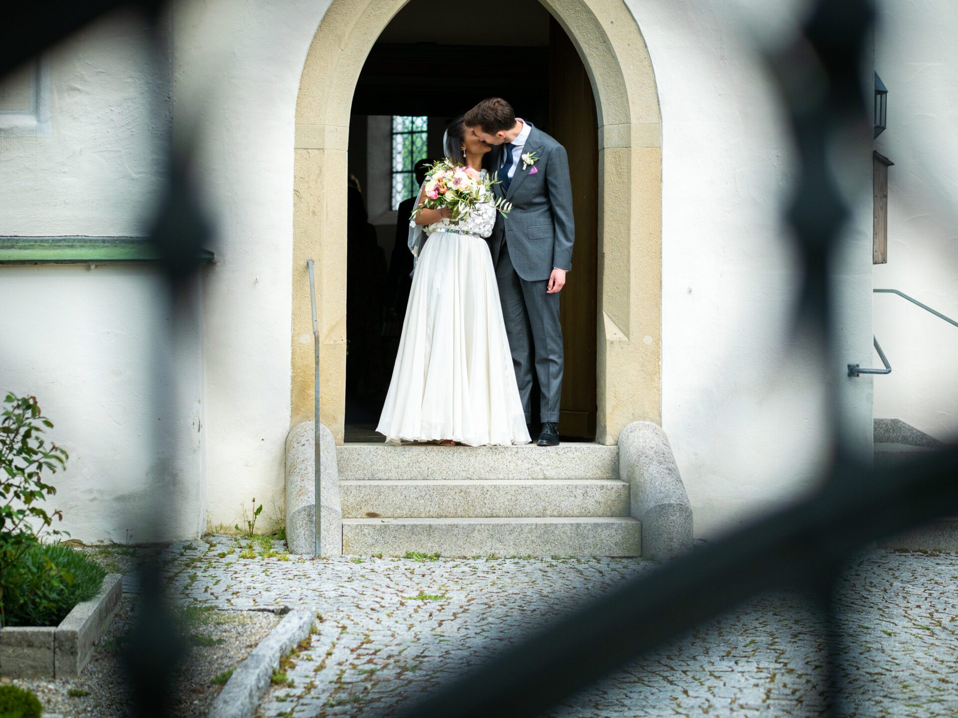 Eine Braut und ein Bräutigam stehen in einer Türöffnung, umarmen sich und lächeln sich an. Aufgenommen von einem Hochzeitsfotografen in München, hält die Braut einen Blumenstrauß, und beide sind in Hochzeitskleidung, schön eingerahmt durch ein dekoratives Tor im Freien. Hochzeitsfotos_Hochzeitsfotograf_München