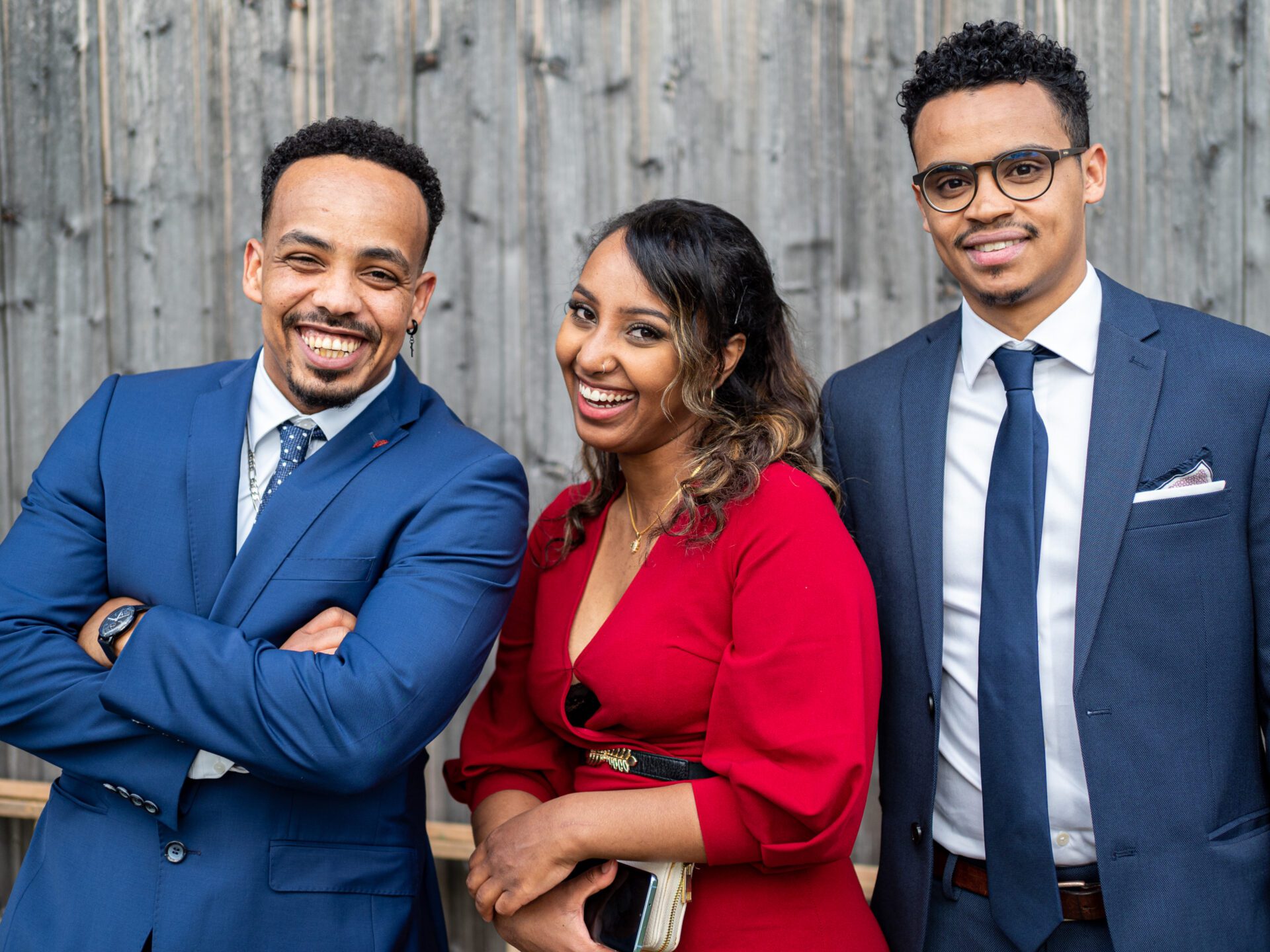 Drei förmlich gekleidete Personen, zwei Männer in blauen Anzügen und eine Frau in einem roten Kleid, stehen lächelnd zusammen vor einem Holzzaun – perfekt für Hochzeitsfotos von einem Hochzeitsfotografen in München. Hochzeitsfotos_Hochzeitsfotograf_München