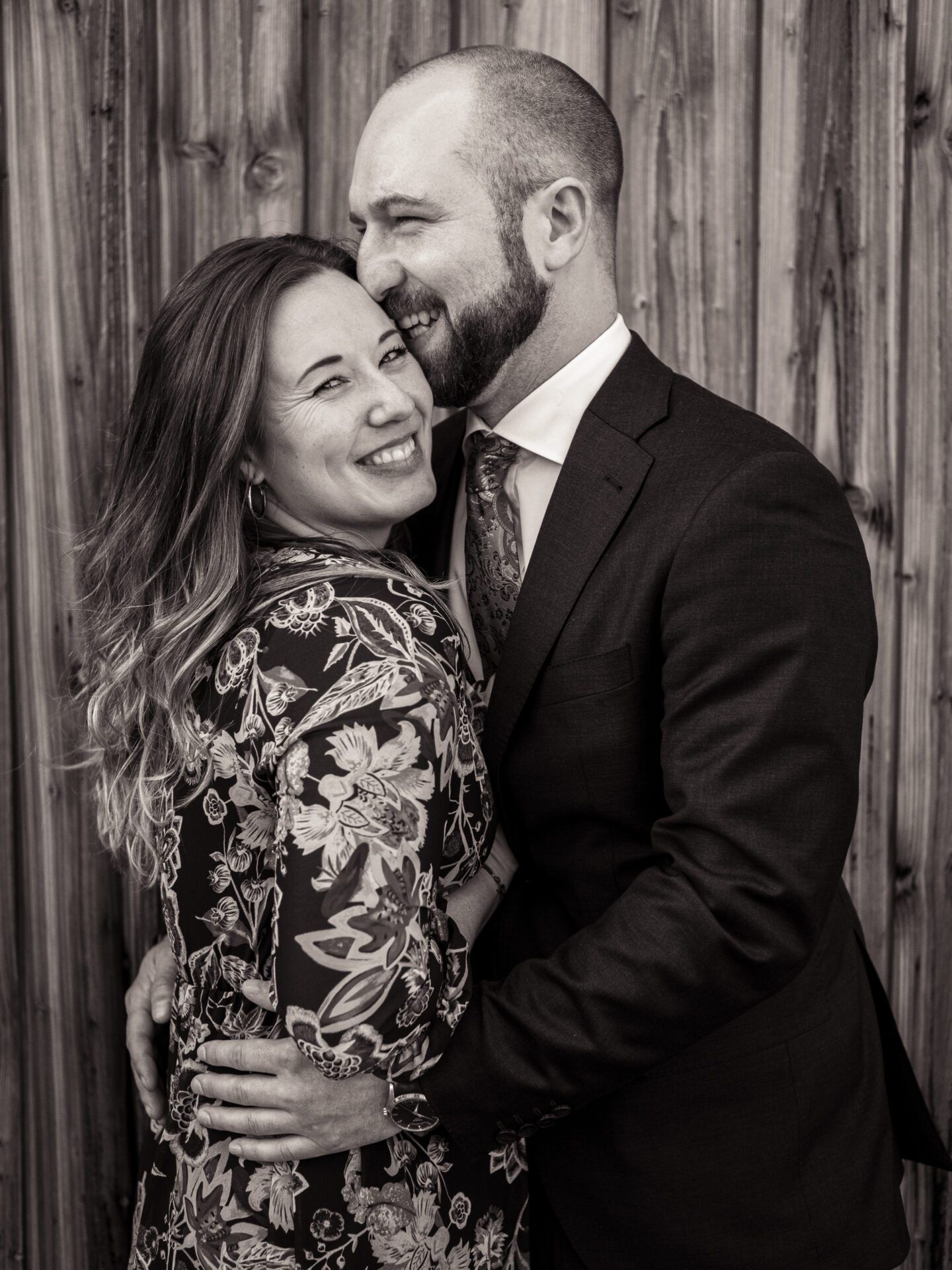 Ein Mann im Anzug umarmt eine lächelnde Frau in einem geblümten Kleid. Sie stehen dicht beieinander vor einer Holzwand und teilen einen fröhlichen und intimen Moment – perfekt für unvergessliche Hochzeitsfotos vom Hochzeitsfotograf aus München. Das Foto ist in schwarz-weiß gehalten. Hochzeitsfotos_Hochzeitsfotograf_München