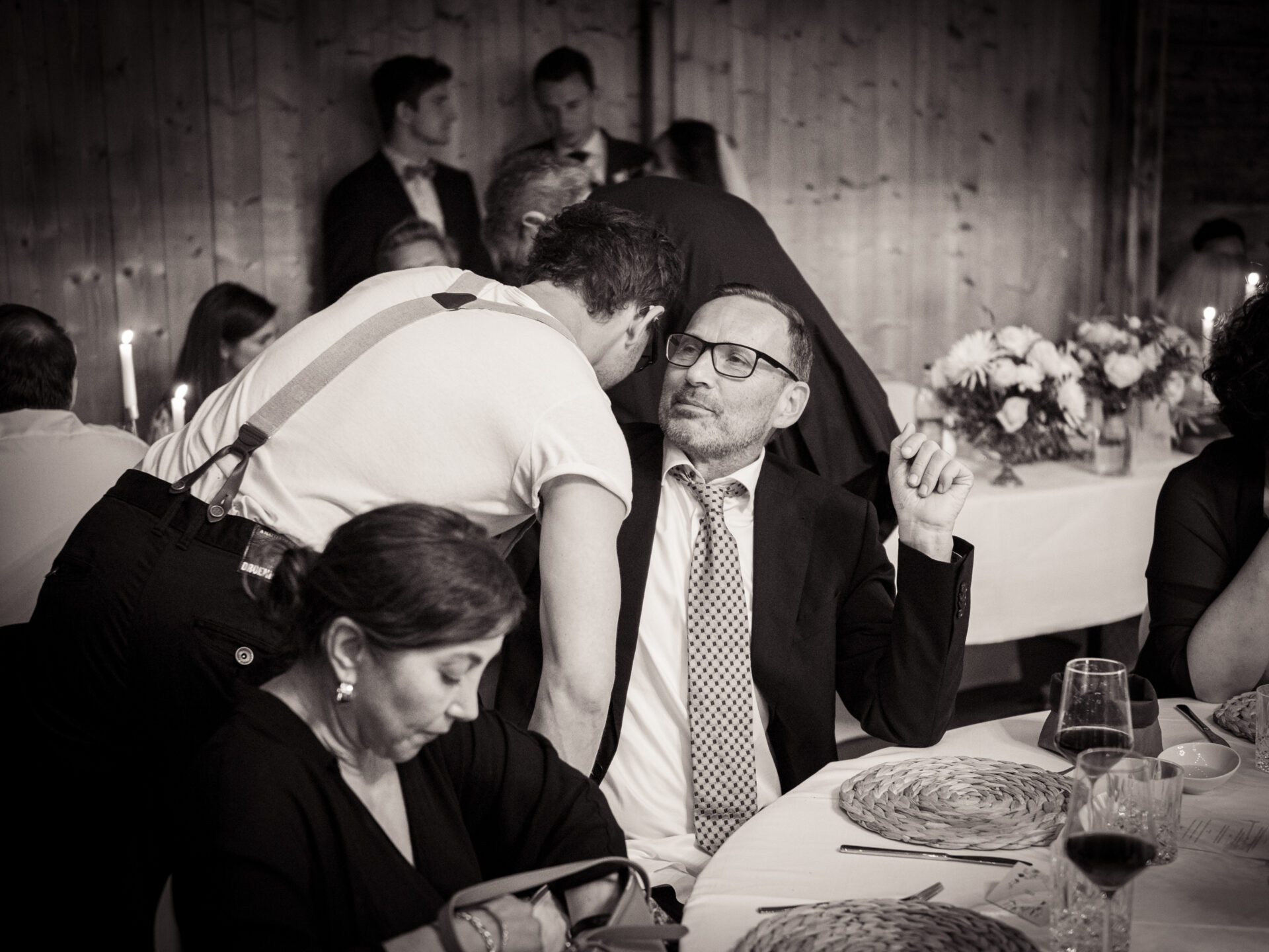 Bei einer formellen Veranstaltung in München beugt sich ein Mann in Hosenträgern vor, um mit einem Gast im Anzug zu sprechen, während eine Frau in der Nähe nach unten schaut. Elegant gedeckte Tische und Blumenarrangements bilden die Kulisse, die ein Hochzeitsfotograf inmitten der sich unter die Gäste mischenden Gäste wunderschön eingefangen hat. Hochzeitsfotos_Hochzeitsfotograf_München