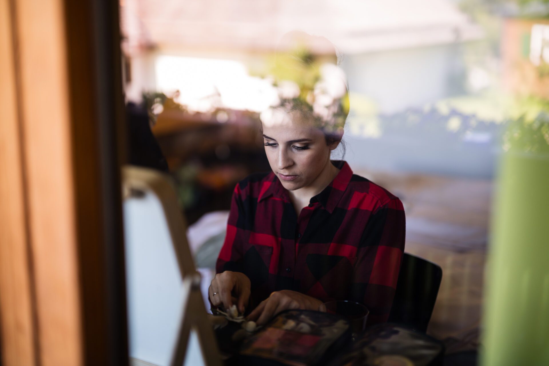 Eine Frau in einem rot-schwarz karierten Hemd sitzt an einem Tisch und schminkt sich. Das Foto, das von einem Hochzeitsfotografen in München aufgenommen wurde, fängt sie durch ein Fenster mit Spiegelungen der Außenszene ein. Hochzeitsfotos_Hochzeitsfotograf_München