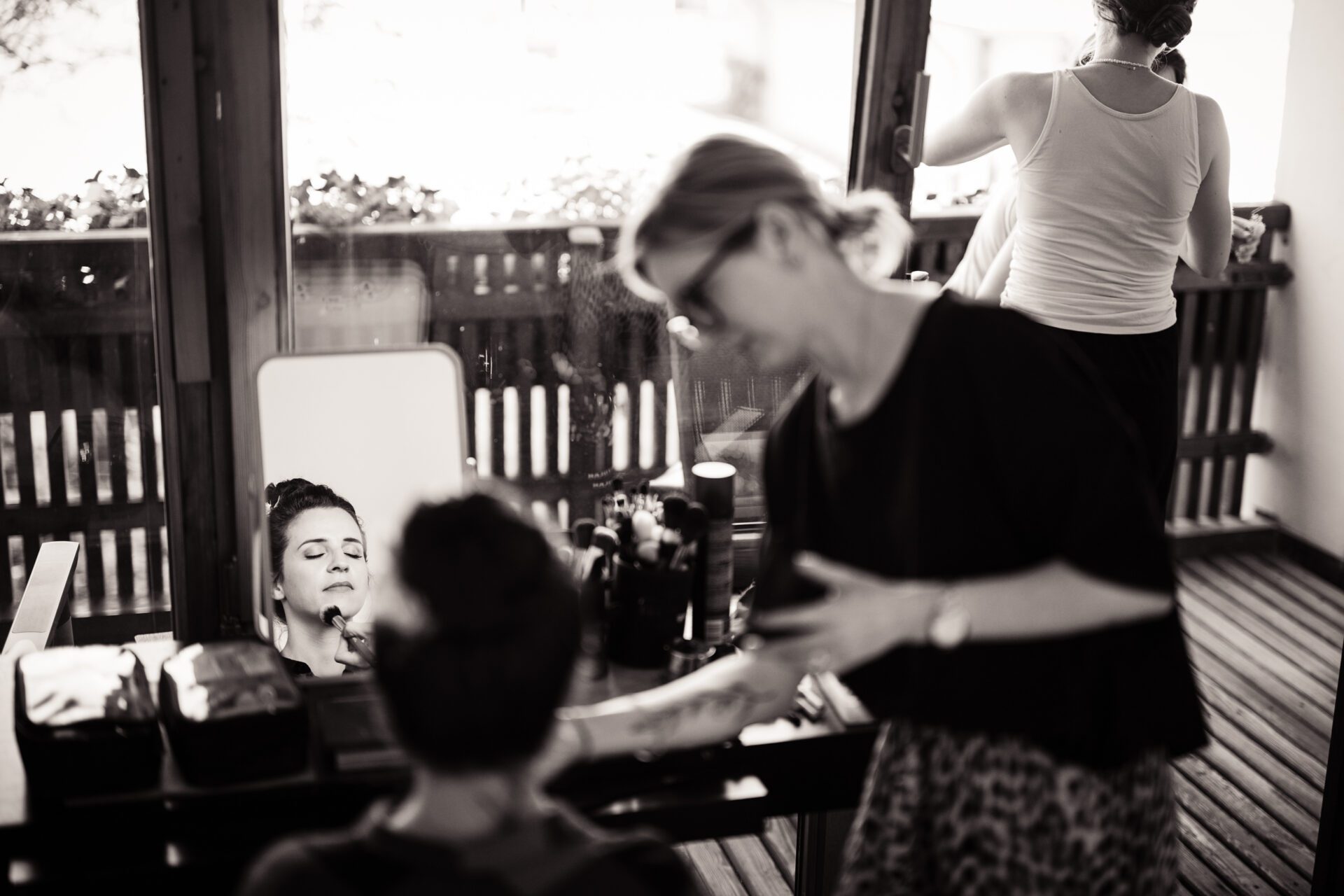 Eine Frau lässt sich schminken, während sie in einen Spiegel schaut, Schminkutensilien liegen auf dem Tisch. Eine andere Frau steht in der Nähe, und eine dritte Person ist am Fenster im Hintergrund zu sehen. Diese schwarz-weiße Szene fängt authentische Hochzeitsfotos in München ein. Hochzeitsfotos_Hochzeitsfotograf_München