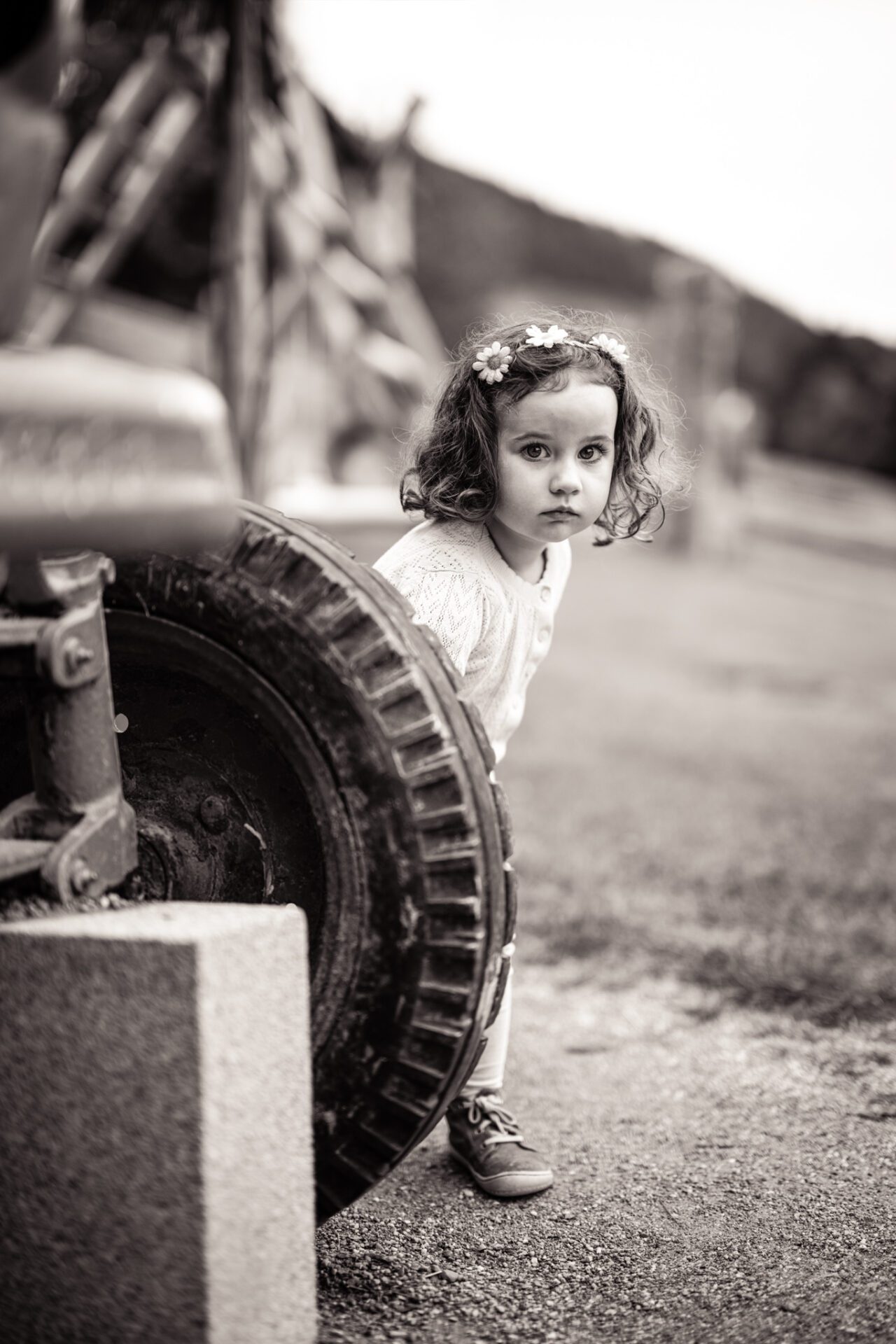 Schwarz-Weiß-Porträt eines kleinen Kindes mit Blumen im lockigen Haar, das neugierig hinter einem großen Traktorreifen hervorschaut.