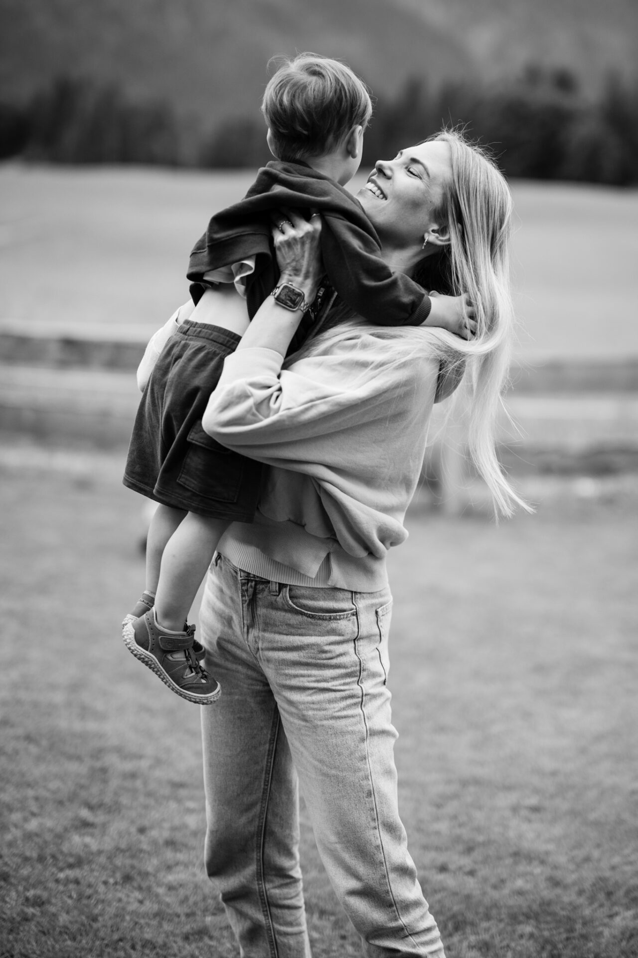 Authentische Familienfotografie in den Bergen: Eine Mutter hebt ihr Kind lachend hoch. Professionelles Schwarz-Weiß-Porträt im Reportagestil.