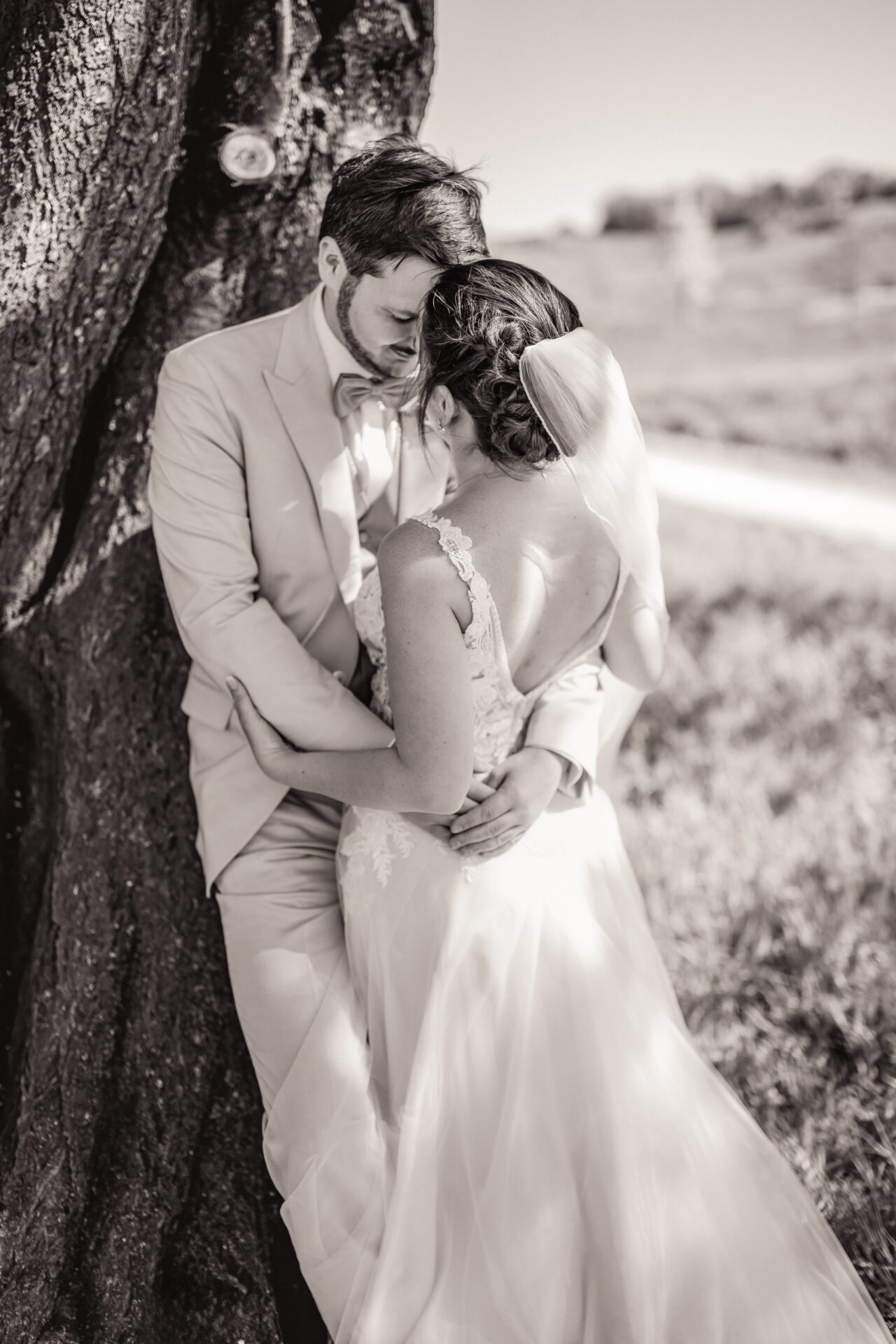 Ein Mann im Anzug umarmt eine lächelnde Frau in einem geblümten Kleid. Sie stehen dicht beieinander vor einer Holzwand und teilen einen fröhlichen und intimen Moment – perfekt für unvergessliche Hochzeitsfotos vom Hochzeitsfotograf aus München. Das Foto ist in schwarz-weiß gehalten. Hochzeitsfotos_Hochzeitsfotograf_München