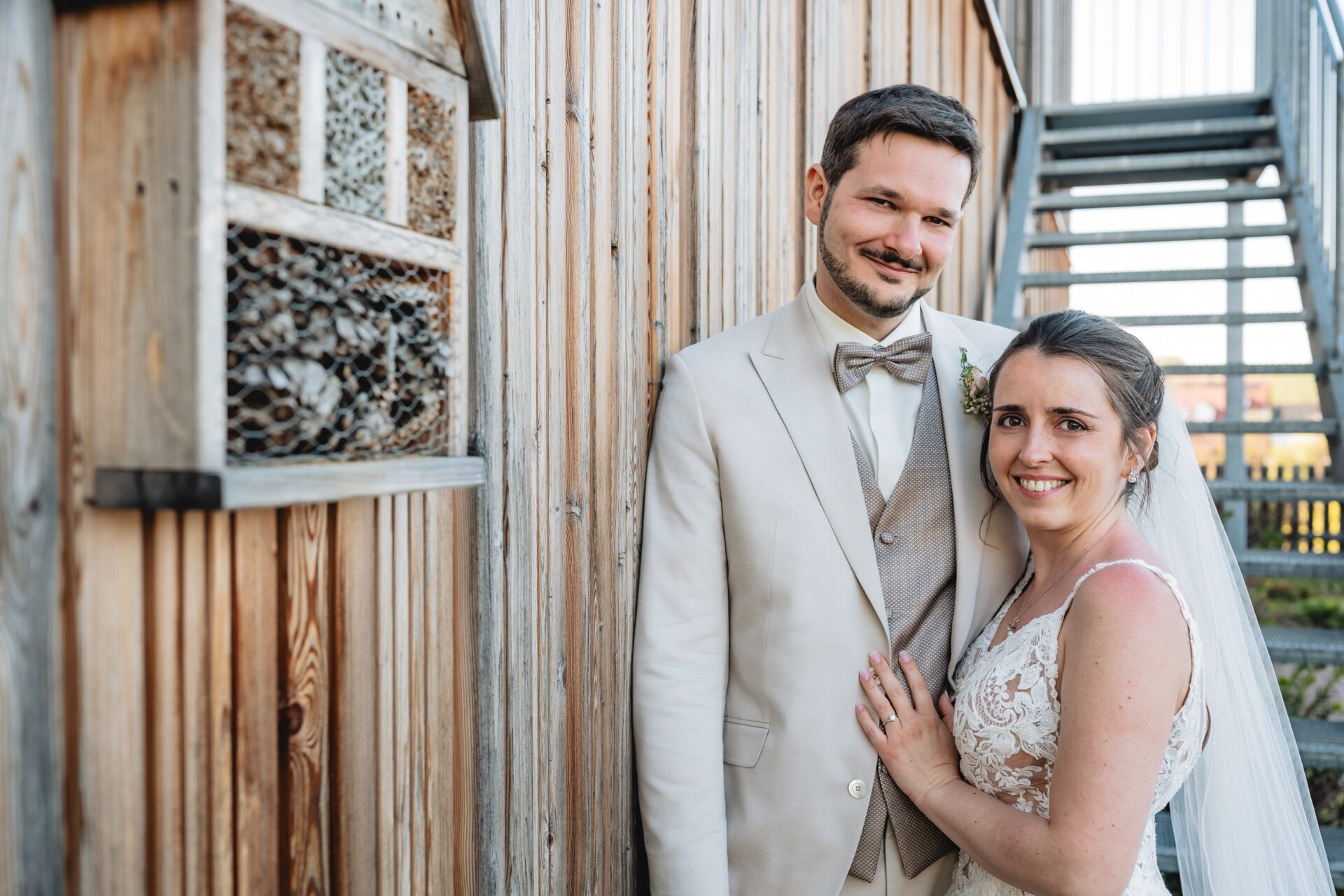 Drei förmlich gekleidete Personen, zwei Männer in blauen Anzügen und eine Frau in einem roten Kleid, stehen lächelnd zusammen vor einem Holzzaun – perfekt für Hochzeitsfotos von einem Hochzeitsfotografen in München. Hochzeitsfotos_Hochzeitsfotograf_München
